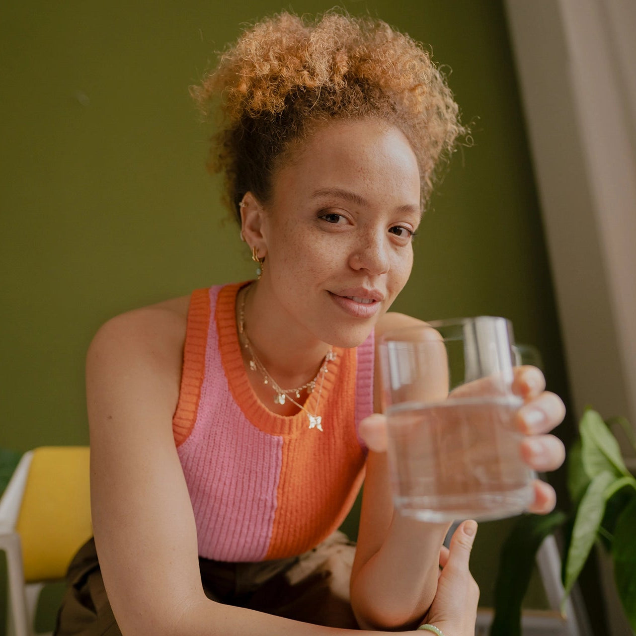 Een vrouw in een roze topje die een glas water vasthoudt, in een huiselijke omgeving met planten.