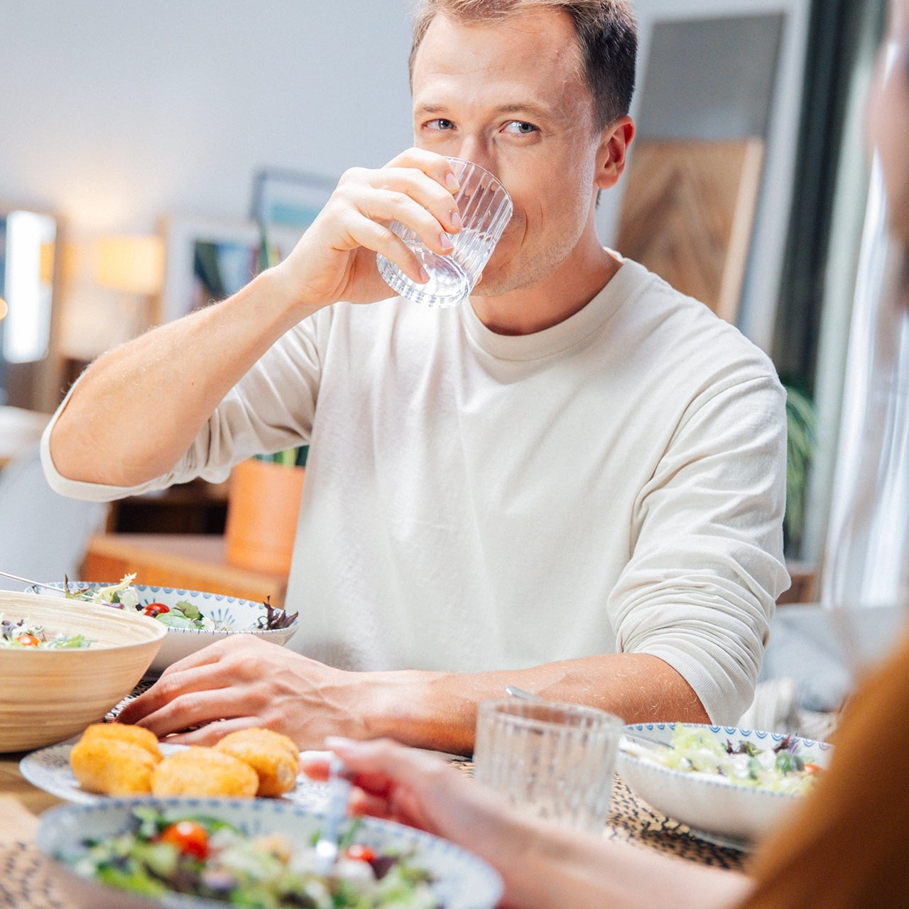 Een man die water drinkt aan een eettafel met eten erop, terwijl een andere persoon gedeeltelijk in beeld is
