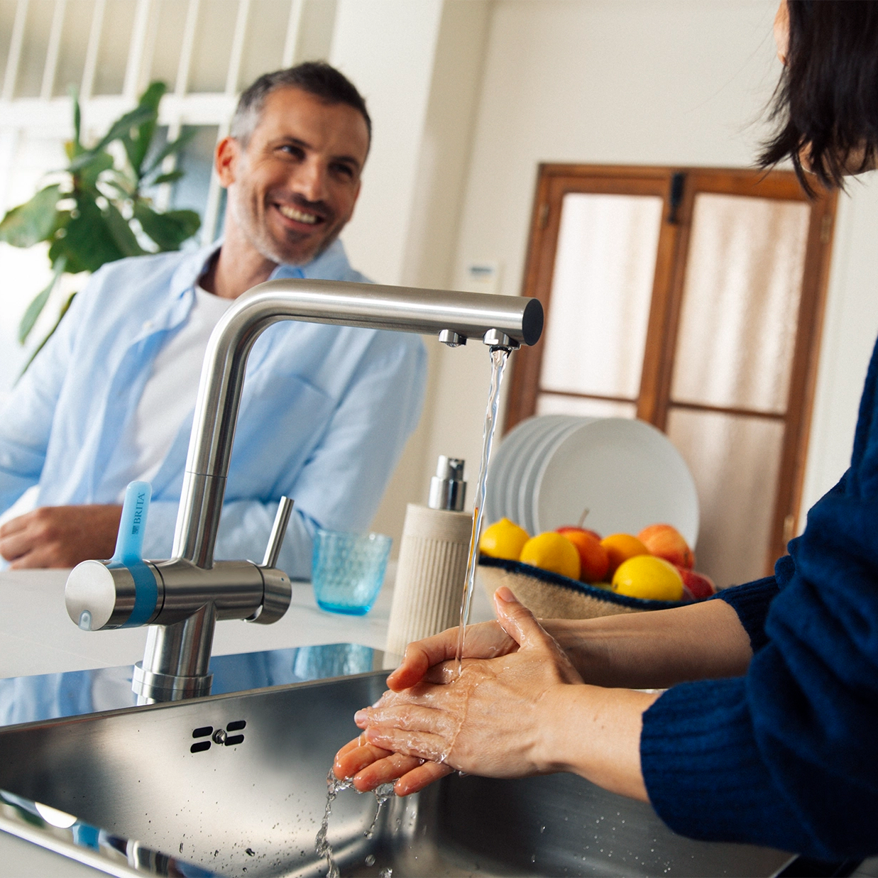 Person washing hands at a kitchen sink with a BRITA 3-way-tap, with a man in the background and fresh fruit on the countertop.
